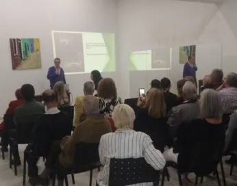 Público sentado sigue una charla frente a una proyección, en una sala con cuadros expuestos.