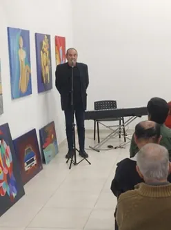 Un orador habla de pie ante el público en una sala de exposición de pinturas.