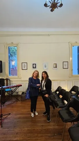 Dos mujeres conversan en un salón con pequeños cuadros enmarcados en la pared.